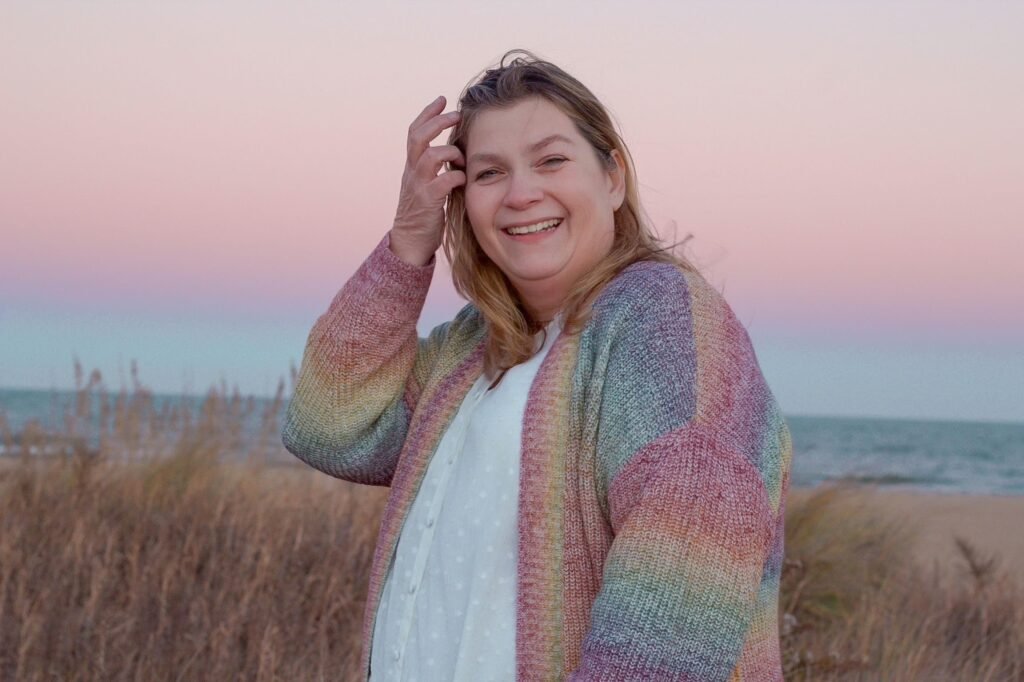 Angie Webber smiling in a pastel rainbow cardigan, standing in beach grass at sunset with the ocean behind her.